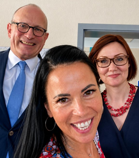 CF-SP_UK Drei Personen stehen in einem Raum und schauen in die Kamera. Eine Frau mit langen, dunklen Haaren und einem bunten Oberteil lächelt. Hinter ihr stehen zwei Personen, die Leiterin der Geschäftsstelle Frau Susanne Pfeiffer mit einer mit Brille und in einem blauen Oberteil, die andere Person mit einem Hemd und Krawatte ist der Studienleiter Prof. Fischer, die Personen lächeln ebenfalls.