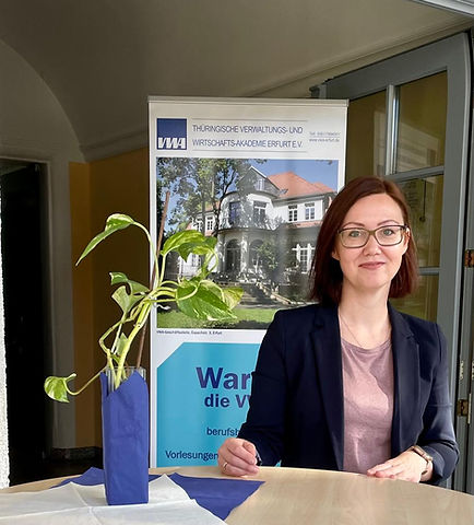 Eine Frau mit braunen, schulterlangen Haaren und Brille sitzt an einem Tisch. Neben ihr steht eine Pflanze in einem Vasenbehälter. Im Hintergrund ist ein Plakat der Thüringischen Verwaltungs- und Wirtschaftsakademie zu sehen, das Informationen zur beruflichen Weiterbildung bietet.